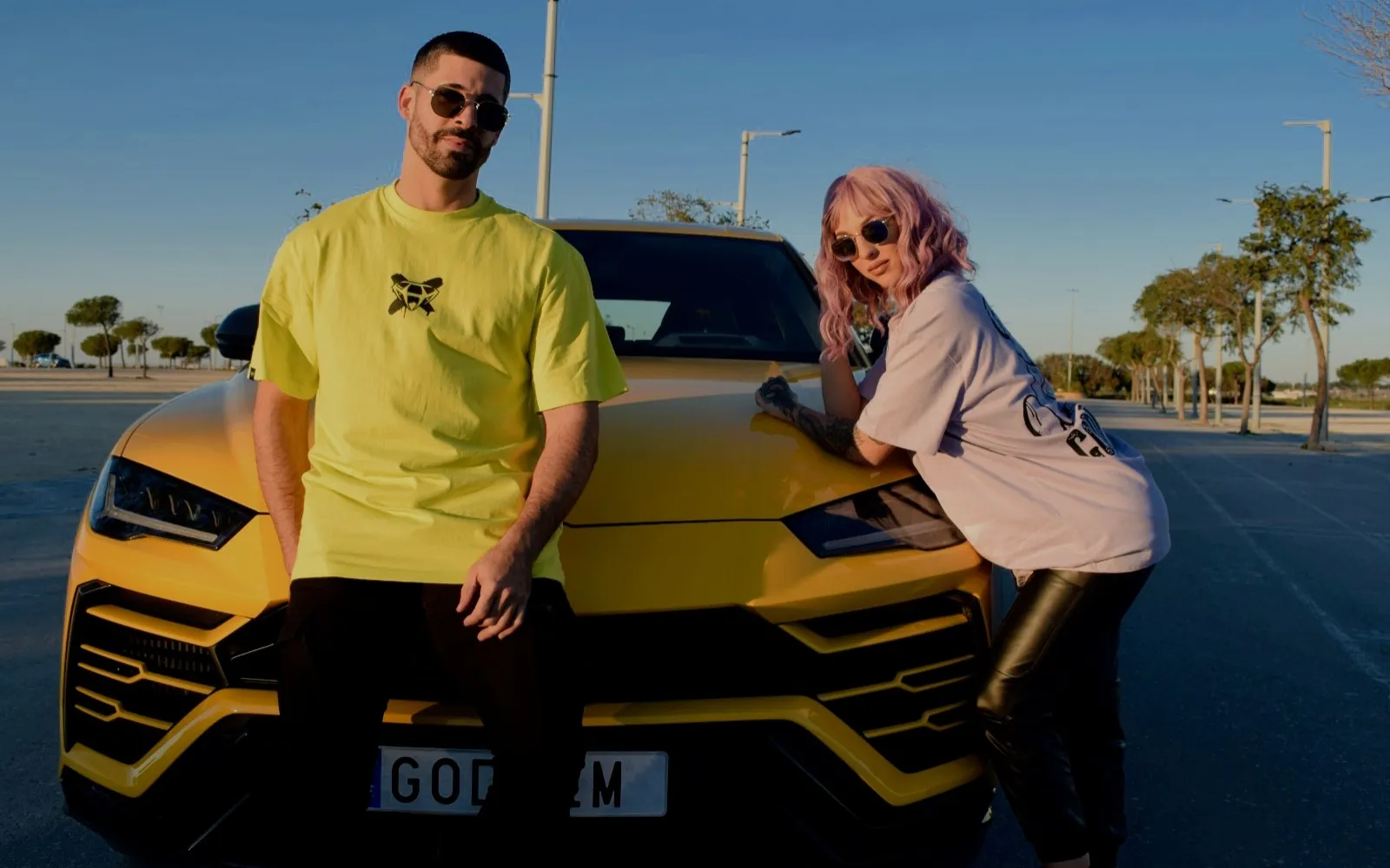 Dos modelos con las camisetas del osito GODHEM en lima y lavanda, apoyados en un coche amarillo
